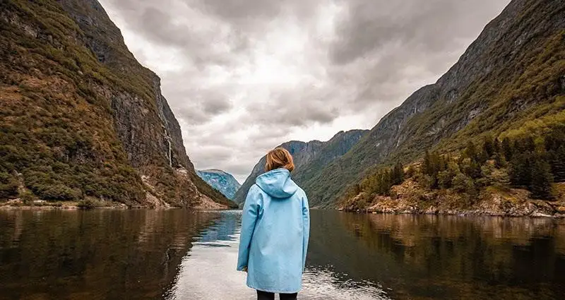 Woman in a blue raincoat stands by a mountain lake in the fall.