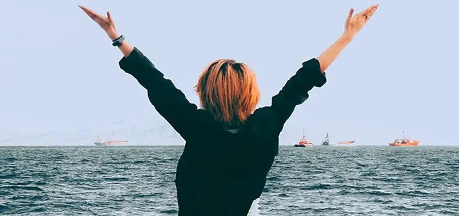 Woman stands near the ocean with hands raised in excitement