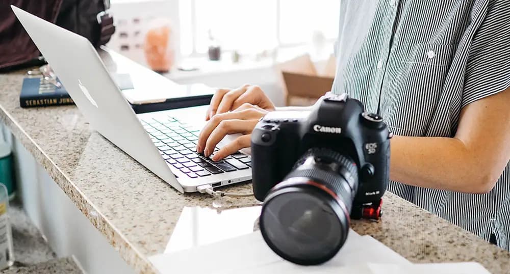 Person types on a laptop with a camera sitting next to them on the counter